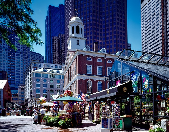 A colonial-era building of red brick with a white cupola sits nestled among skyscrapers. In the foreground is a glass building with a patio table in front of it. 
