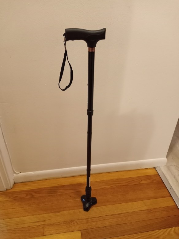 A tripod cane stands alone on a wood floor, its strap dangling from the handle.