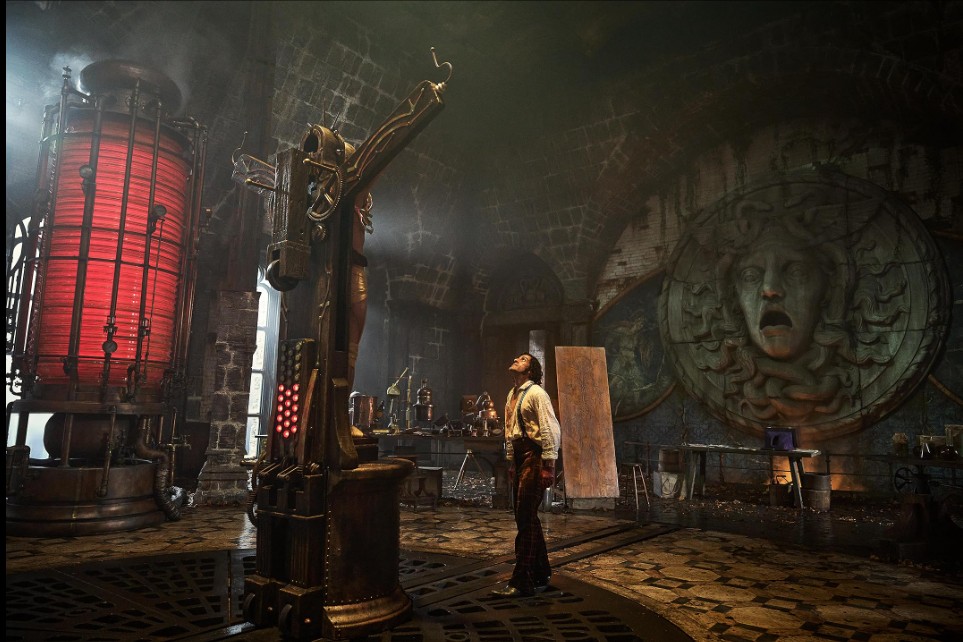Victor Frankenstein stands beneath a cruciform apparatus for bringing his creation to life. The room is circular, dark, and imposing, with a massive stone carving on the back wall and at left, a huge metal and red glass cylinder - the battery by which he will harness the lightning's energy. 
