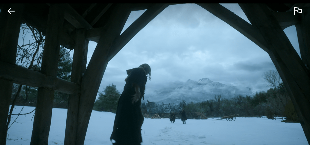 A wounded Adam stands outside under a cloudy sky near the gatehouse beyond the old man's cottage. Two hunters are advancing toward him, guns raised. 